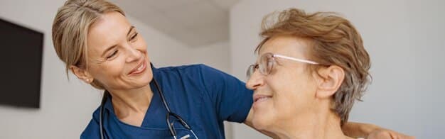 An eye-level shot of a caring nurse assisting a senior patient in a German care facility. Both are smiling and engaged in a conversation. The environment looks comfortable and home-like, designed to convey quality elder care.
